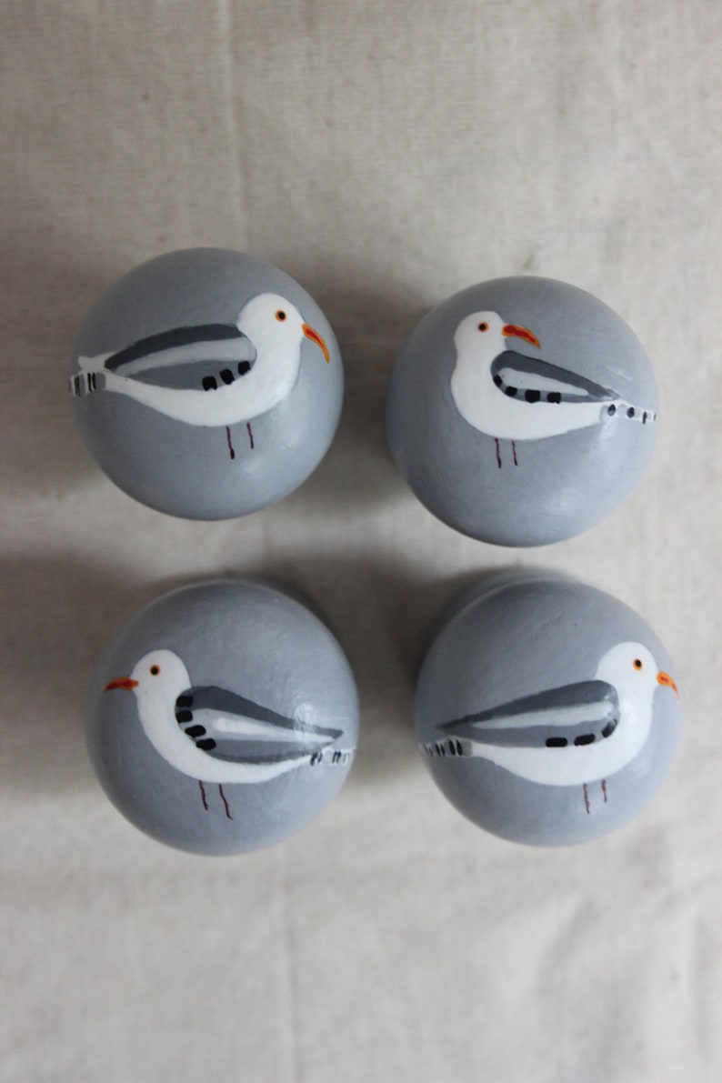 K&ouml;nnte beinhalten: Vier graue runde Kn&ouml;pfe mit bemalten M&ouml;wenmotiven. Jeder Knopf zeigt eine wei&szlig;e M&ouml;we mit schwarzen Markierungen und einem orangefarbenen Schnabel.