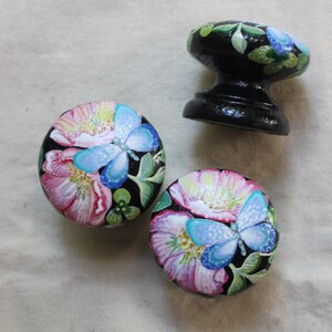 May include: Three black wooden knobs with a floral design. Each knob features a pink and white flower with a blue butterfly on a black background.