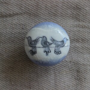 May include: A spherical decorative item, white with a light blue rim, featuring three grey birds on a wire. The birds are hand-painted with black detailing. The object rests on a textured, neutral-toned surface.