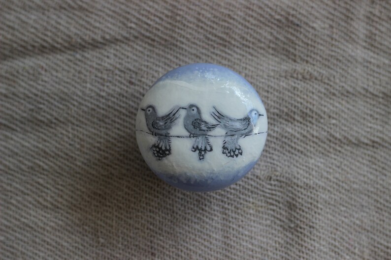 May include: A spherical decorative item, white with a light blue rim, featuring three grey birds on a wire. The birds are hand-painted with black detailing. The object rests on a textured, neutral-toned surface.
