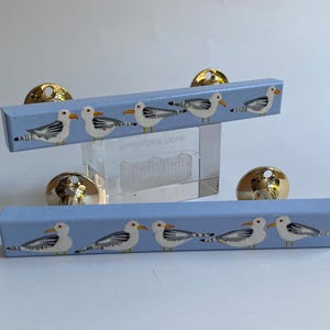 Könnte beinhalten: Zwei hellblaue Schrankgriffe mit goldfarbenen Knöpfen. Jeder Griff ist mit handgemalten Möwen verziert. Die Griffe sind rechteckig und auf einem transparenten Acrylblock montiert.