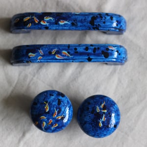 May include: A set of blue cabinet hardware, including two handles and two knobs. The hardware is decorated with a koi fish design in orange, yellow, and white, with black accents. The hardware is likely made of ceramic.
