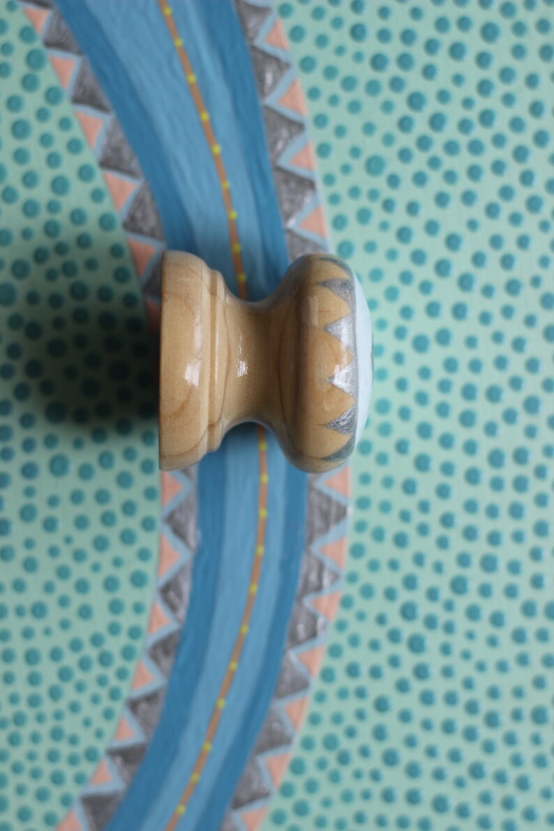 Puede incluir: Un tirador de armario de madera clara, rematado con un dise&ntilde;o plateado y blanco. El tirador se encuentra sobre un fondo de dise&ntilde;o de r&iacute;o azul con un fondo turquesa punteado.