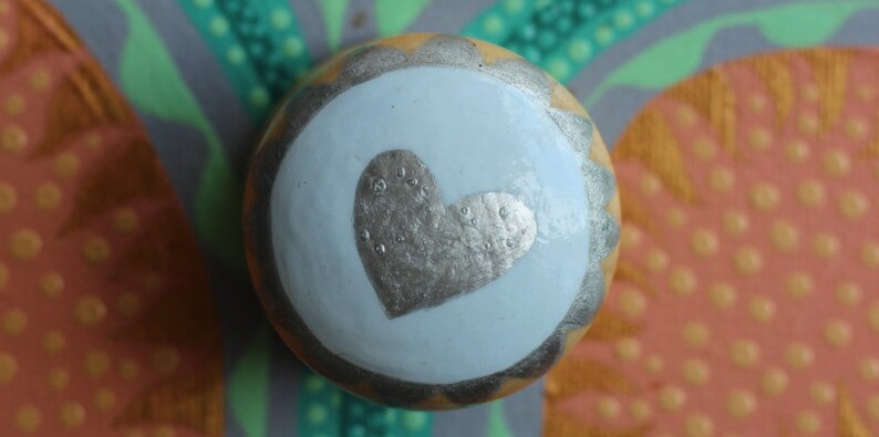 Pu&ograve; includere: Un pomello decorativo rotondo con un centro azzurro con un disegno a cuore argentato. Il pomello ha un bordo argentato smerlato e un bordo esterno giallo-arancio. Lo sfondo presenta un motivo astratto colorato.