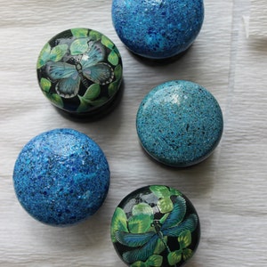 Op de afbeelding: Een verzameling decoratieve ronde objecten. Sommige zijn effen blauw met een getextureerde afwerking, terwijl andere vlinder- en bladmotieven in blauw en groen op een zwarte achtergrond hebben. De objecten zijn gerangschikt op een wit oppervlak.