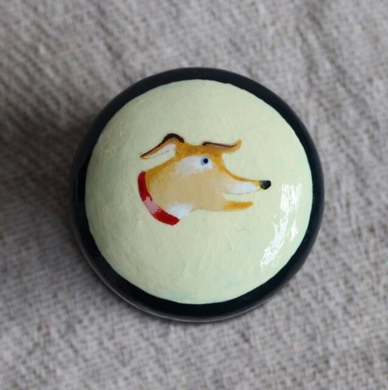May include: A circular, painted item with a dog's head design. The dog has tan fur, a red collar, and blue eyes, set against a pale yellow backdrop. The object is framed by a black border.