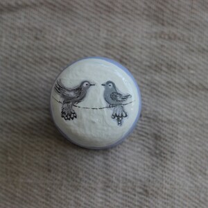 May include: A round, hand-painted decorative knob with two grey birds on a black line against a white and pale blue background. The birds face each other, creating a charming, whimsical design.