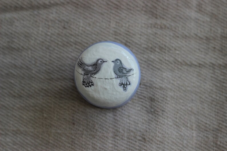 May include: A round, hand-painted decorative knob with two grey birds on a black line against a white and pale blue background. The birds face each other, creating a charming, whimsical design.