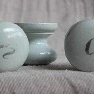 May include: Three light blue ceramic knobs with silver lettering. Two are round with a letter each, and one is a cylindrical shape. The knobs are on a neutral-colored fabric.