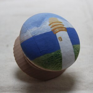 May include: A wooden bottle stopper featuring a hand-painted lighthouse scene. The design includes a white lighthouse with a tan top, set against a blue sky and sea, and green grass. The stopper is made of light brown wood.
