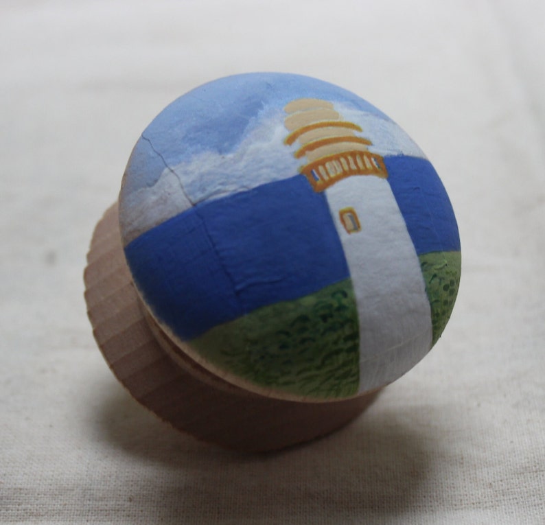 May include: A wooden bottle stopper featuring a hand-painted lighthouse scene. The design includes a white lighthouse with a tan top, set against a blue sky and sea, and green grass. The stopper is made of light brown wood.