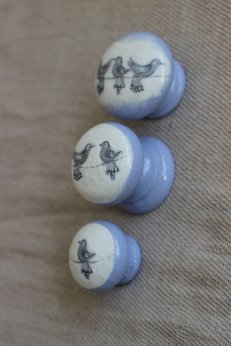 May include: Three pale blue cabinet knobs with a white top featuring a black and grey bird design. The knobs are arranged vertically, showcasing the bird illustrations on each knob. The background is a neutral fabric.