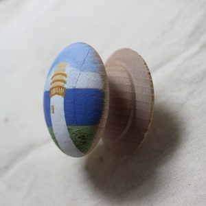 May include: A wooden cabinet knob with a painted lighthouse scene. The knob features a white lighthouse with a yellow top, set against a blue sky and sea, and green grass. The unpainted wooden base is visible.