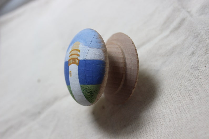 May include: A wooden cabinet knob with a painted lighthouse scene. The knob features a white lighthouse with a yellow top, set against a blue sky and sea, and green grass. The unpainted wooden base is visible.