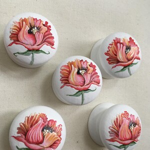 Puede incluir: Cinco pomos de armario blancos con dise&ntilde;os florales pintados a mano. Cada pomo presenta una vibrante flor roja y naranja con tallos y hojas verdes. Los pomos son redondos y tienen un acabado liso.