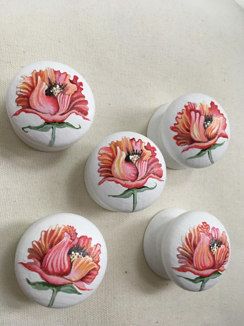 Puede incluir: Cinco pomos de armario blancos con dise&ntilde;os florales pintados a mano. Cada pomo presenta una vibrante flor roja y naranja con tallos y hojas verdes. Los pomos son redondos y tienen un acabado liso.