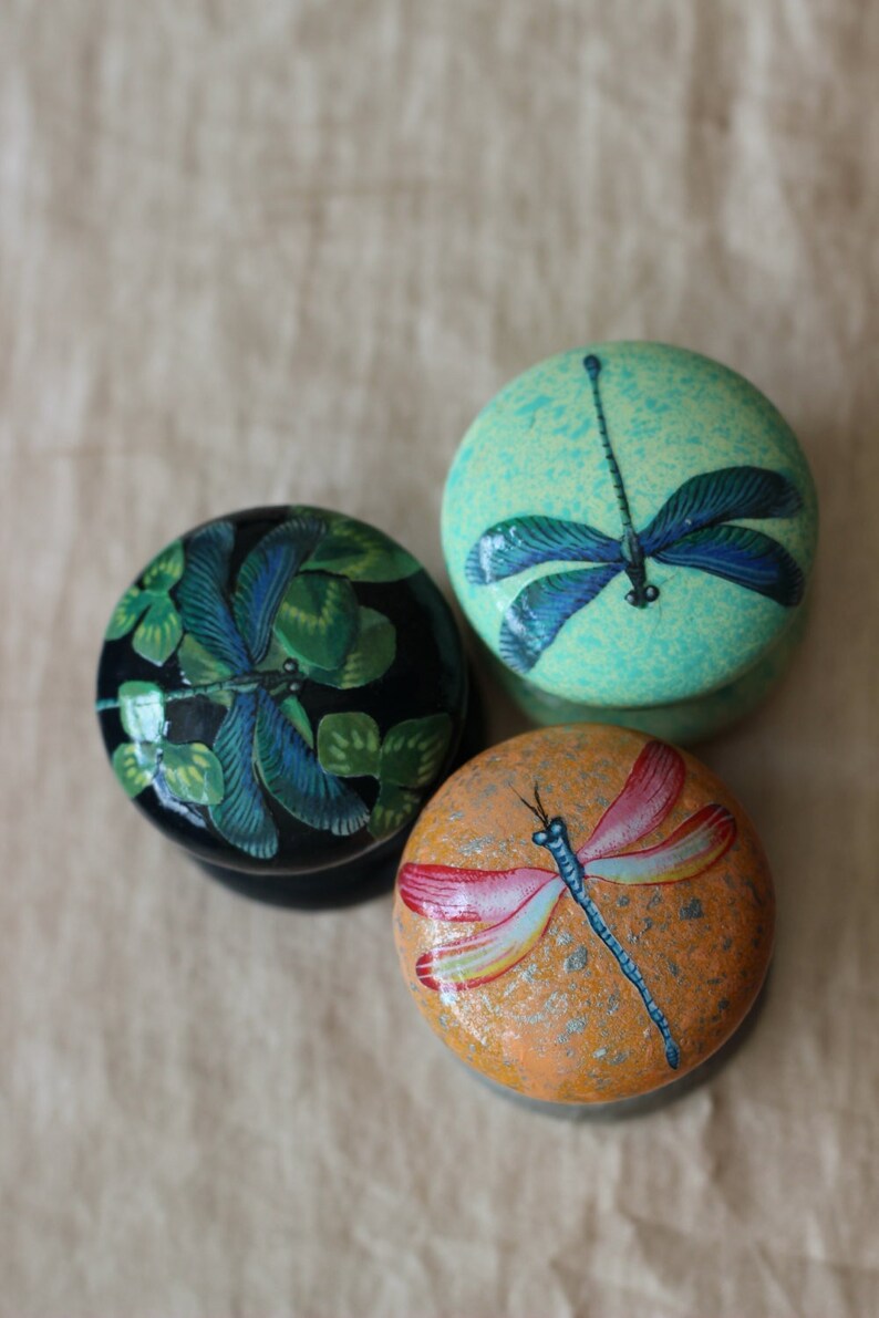 Może przedstawiać: Trzy okrągłe drewniane ozdoby z malowanymi wzorami ważek. Ozdoby są pomalowane w odcieniach zieleni, błękitu i pomarańczy.