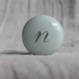 Puede incluir: Un pomo redondo de color azul claro con la letra "n" escrita en plata en la parte delantera.