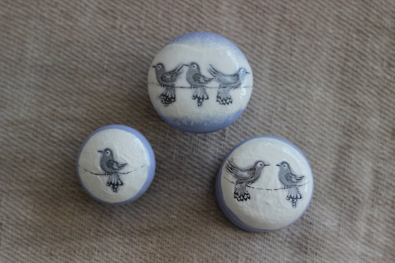 May include: Three round decorative objects with a white base and a light blue rim. Each object features a hand-painted design of birds perched on a wire. The birds are depicted in shades of grey and black.