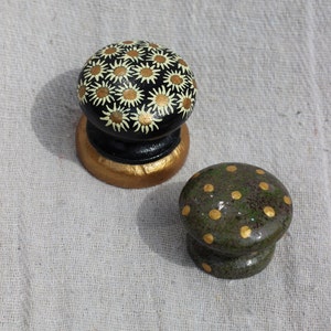 May include: Two decorative wooden knobs, one with a black background and white daisy pattern, the other with a green background and gold polka dots.