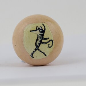 Pode incluir: Puxador de madeira redondo, cor p&ecirc;ssego claro, com uma ilustra&ccedil;&atilde;o pintada &agrave; m&atilde;o de uma criatura estilizada dan&ccedil;ando. A criatura &eacute; preta e branca com uma cauda longa, sobre um fundo amarelo p&aacute;lido.