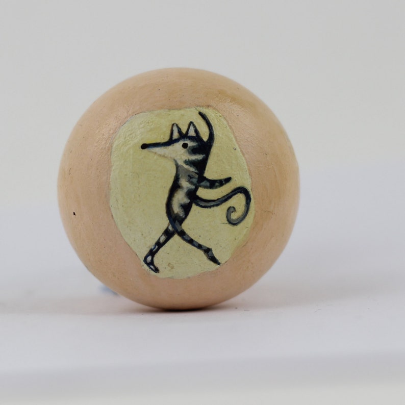 Pode incluir: Puxador de madeira redondo, cor p&ecirc;ssego claro, com uma ilustra&ccedil;&atilde;o pintada &agrave; m&atilde;o de uma criatura estilizada dan&ccedil;ando. A criatura &eacute; preta e branca com uma cauda longa, sobre um fundo amarelo p&aacute;lido.