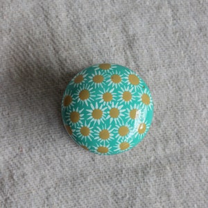 Pode incluir: Uma pedra pintada de turquesa com um padr&atilde;o de margaridas brancas e douradas.
