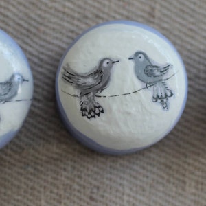 May include: Three round decorative buttons with a bird design. The buttons are white with a pale blue border. Each button features a different number of grey birds perched on a line.