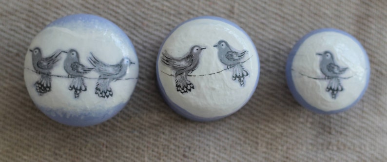 May include: Three round decorative buttons with a bird design. The buttons are white with a pale blue border. Each button features a different number of grey birds perched on a line.