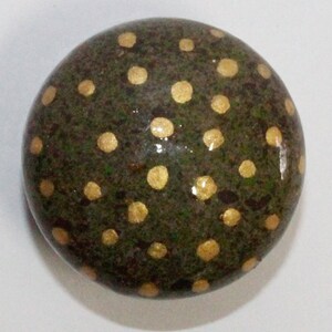 Può includere: Un sasso rotondo di colore verde scuro con pois dorati dipinti sulla superficie.