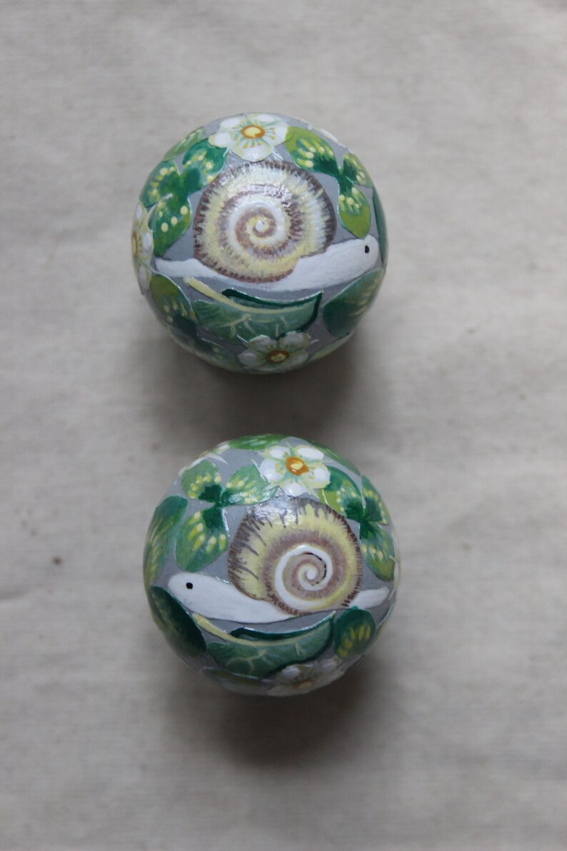 May include: Two decorative, round objects with a gray background. Each object is painted with a snail design, featuring a cream-colored shell, white body, and surrounded by green leaves and white flowers. The overall theme is nature-inspired.