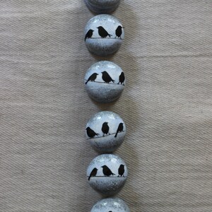 K&ouml;nnte beinhalten: Sechs runde, wei&szlig;e Keramikkn&ouml;pfe mit schwarzen Silhouetten von V&ouml;geln, die auf einem Draht sitzen. Die Kn&ouml;pfe haben ein abgenutztes, antikes Aussehen.