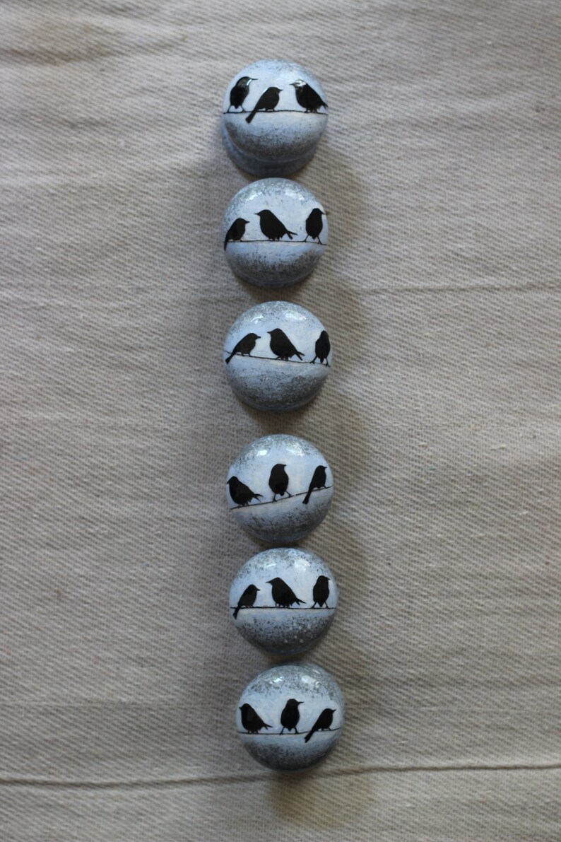 K&ouml;nnte beinhalten: Sechs runde, wei&szlig;e Keramikkn&ouml;pfe mit schwarzen Silhouetten von V&ouml;geln, die auf einem Draht sitzen. Die Kn&ouml;pfe haben ein abgenutztes, antikes Aussehen.