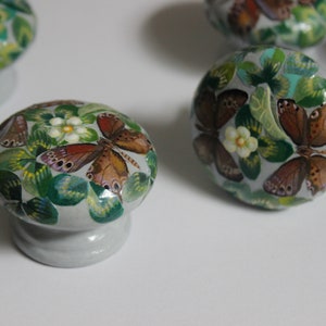 Puede incluir: Cuatro pomos de caj&oacute;n de cer&aacute;mica con una base gris claro y un dise&ntilde;o pintado de hojas verdes, flores blancas y mariposas marrones.