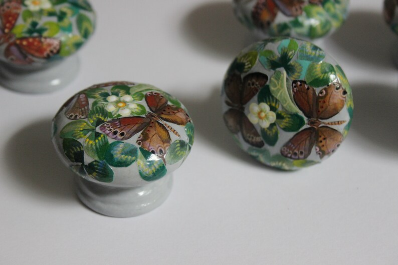 Puede incluir: Cuatro pomos de caj&oacute;n de cer&aacute;mica con una base gris claro y un dise&ntilde;o pintado de hojas verdes, flores blancas y mariposas marrones.