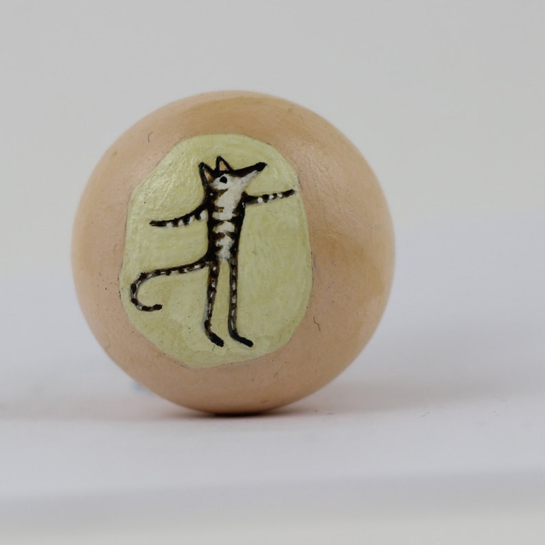 Pode incluir: Objeto decorativo esf&eacute;rico bege com uma ilustra&ccedil;&atilde;o pintada de um animal estilizado com corpo listrado e bra&ccedil;os estendidos. O animal est&aacute; sobre um oval verde claro.