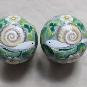 May include: Two round, decorative objects with hand-painted snail and floral designs. The snails have cream-colored shells and white bodies, set against a gray background with green leaves and white flowers. The objects are likely decorative ornaments.