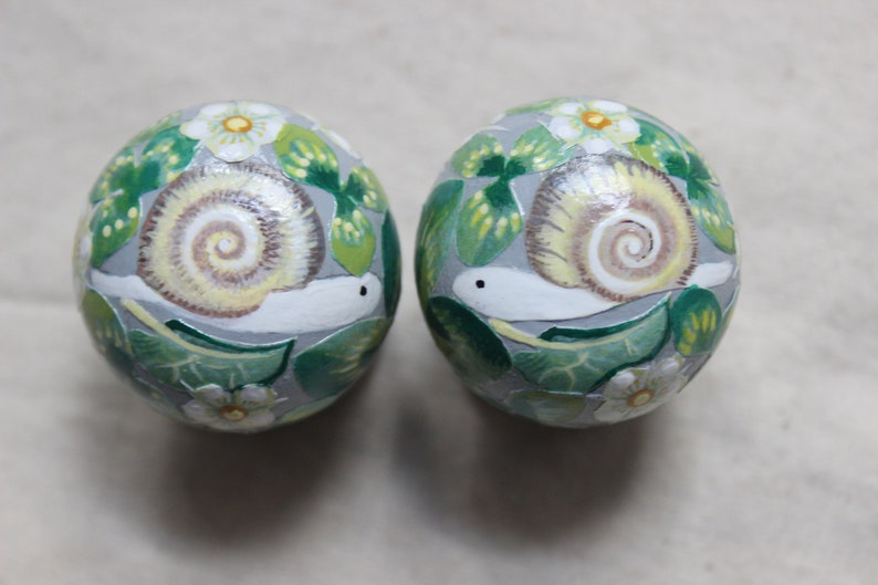 May include: Two round, decorative objects with hand-painted snail and floral designs. The snails have cream-colored shells and white bodies, set against a gray background with green leaves and white flowers. The objects are likely decorative ornaments.