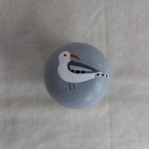 K&ouml;nnte beinhalten: Ein runder, grauer Anh&auml;nger mit einem darauf gemalten wei&szlig;en Seemann. Der Seemann hat einen schwarz-wei&szlig; gestreiften Fl&uuml;gel und einen roten Schnabel.