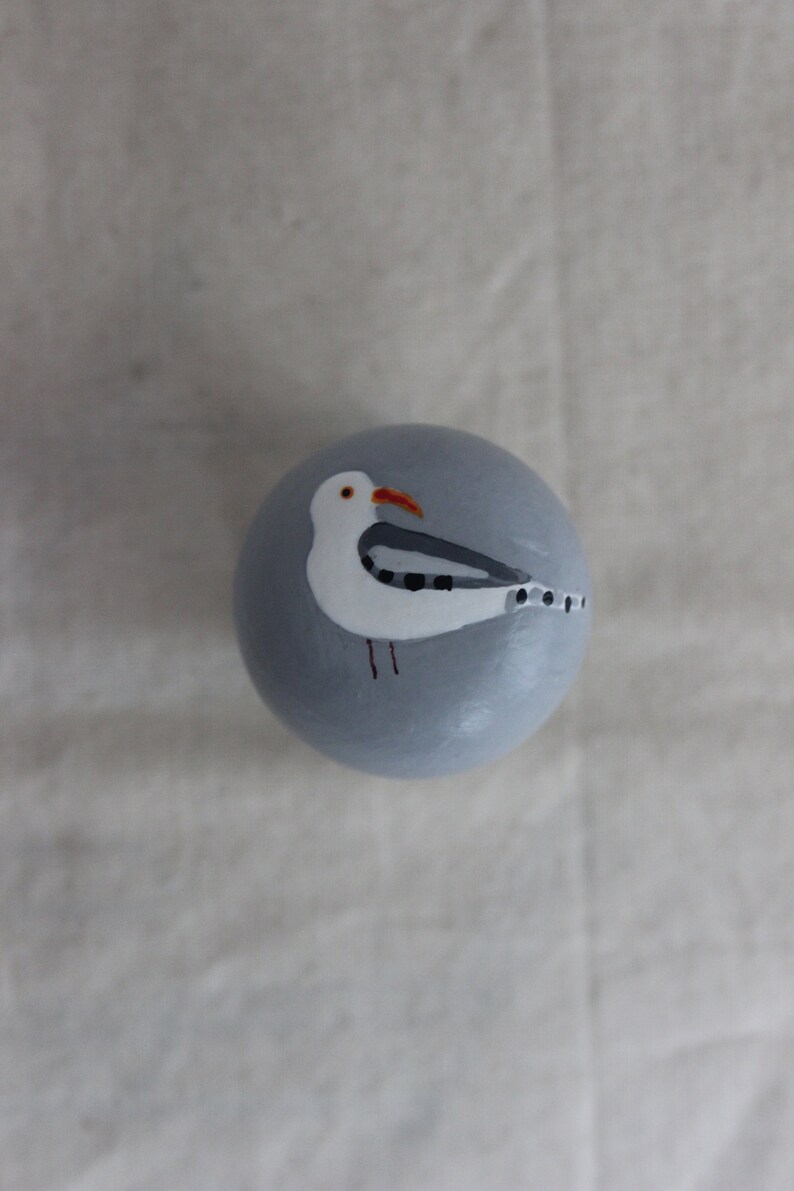 K&ouml;nnte beinhalten: Ein runder, grauer Anh&auml;nger mit einem darauf gemalten wei&szlig;en Seemann. Der Seemann hat einen schwarz-wei&szlig; gestreiften Fl&uuml;gel und einen roten Schnabel.