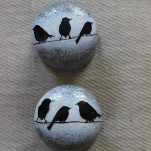 K&ouml;nnte beinhalten: Zwei runde Keramikornamente mit einem blau-grauen, gesprenkelten Hintergrund. Jedes Ornament zeigt eine schwarze Silhouette von V&ouml;geln, die auf einem Draht sitzen.