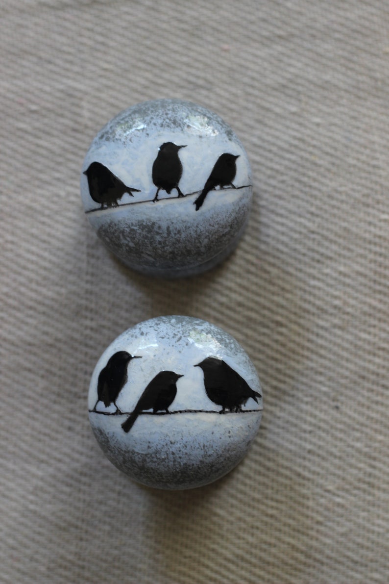 K&ouml;nnte beinhalten: Zwei runde Keramikornamente mit einem blau-grauen, gesprenkelten Hintergrund. Jedes Ornament zeigt eine schwarze Silhouette von V&ouml;geln, die auf einem Draht sitzen.