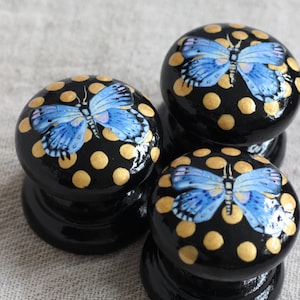 以下が含まれることがあります： 光沢のある黒いキャビネットノブが3つ。各ノブには、金色の水玉模様の青い蝶のデザインが施されています。ノブはおそらく木材または同様の素材で作られており、家具に適しています。