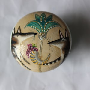 May include: A round, beige decorative object with a painted design featuring two jackals facing each other. A green and white stylized plant, a horn, and grapes are also depicted. The object has a light blue border.