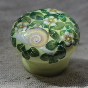 Op de afbeelding: Een kleine, gele keramische knop met een geschilderd ontwerp van groene bladeren, witte bloemen en een slak. De slak is geschilderd in tinten roze, geel en oranje.