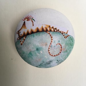 Puede incluir: Una piedra redonda pintada a mano presenta un dise&ntilde;o caprichoso de gato. El gato, con rayas marrones y naranjas, est&aacute; tumbado sobre una superficie verde y azul. Una flor adorna su cabeza. La piedra est&aacute; pintada con t&eacute;cnicas similares a la acuarela.
