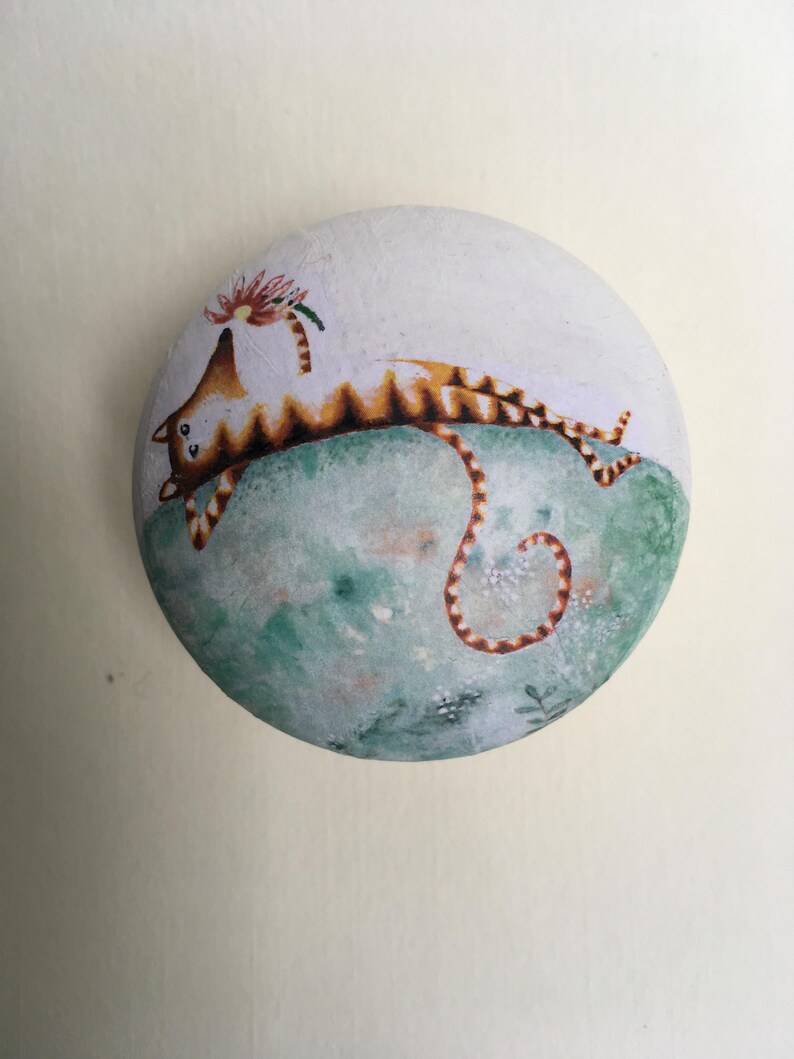 Puede incluir: Una piedra redonda pintada a mano presenta un dise&ntilde;o caprichoso de gato. El gato, con rayas marrones y naranjas, est&aacute; tumbado sobre una superficie verde y azul. Una flor adorna su cabeza. La piedra est&aacute; pintada con t&eacute;cnicas similares a la acuarela.