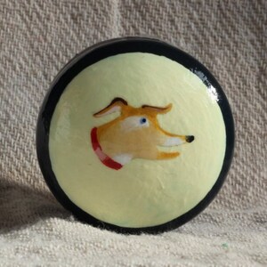 Puede incluir: Perilla decorativa redonda pintada a mano. Presenta un fondo amarillo claro con la cabeza de un perro de dibujos animados de perfil. El perro tiene pelaje marr&oacute;n, ojos azules y un collar rojo. La perilla tiene un borde negro.