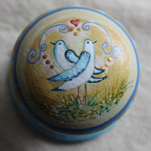 May include: A hand-painted ceramic egg featuring a blue and white design of two birds beneath a heart. The egg is decorated with gold accents and sits on a blue base.