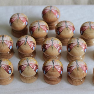 Könnte beinhalten: Eine Sammlung von Holzschrankknöpfen. Jeder Knopf hat ein handgemaltes Schmetterlingsdesign mit roten, blauen und gelben Akzenten. Die Knöpfe sind hellbraun und haben eine abgerundete Oberseite und einen zylindrischen Sockel.